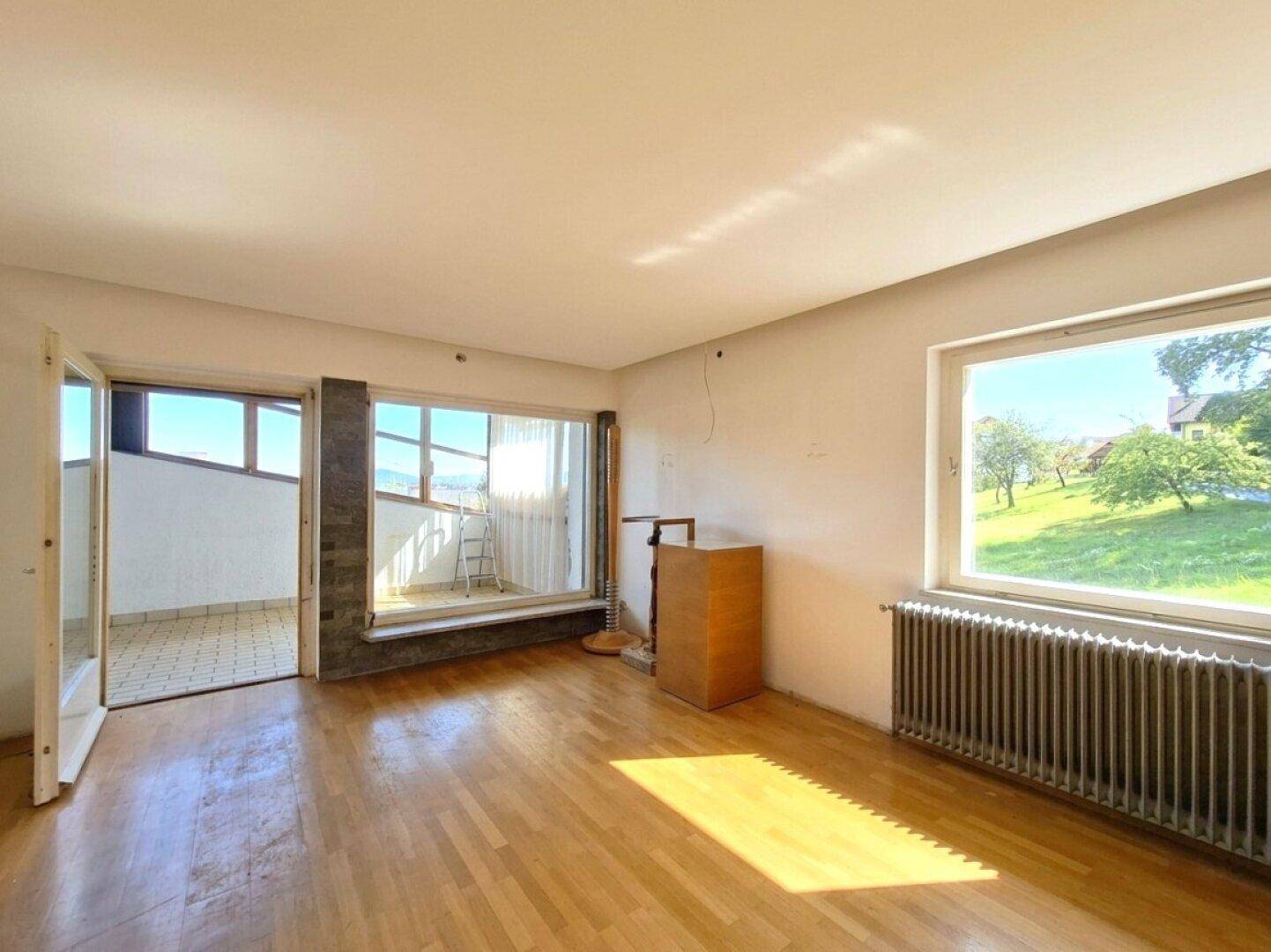 Lichtdurchflutetes Wohnzimmer mit Parkettboden, großem Fenster und Zugang zu einem Außenbereich.
