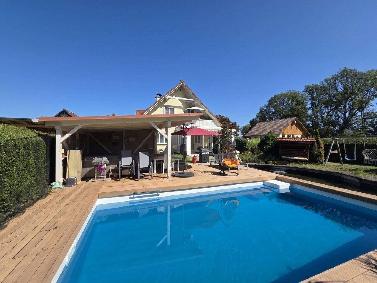 Großer Außenbereich mit Swimmingpool, Holzdeck und überdachtem Sitzbereich vor dem Haus.