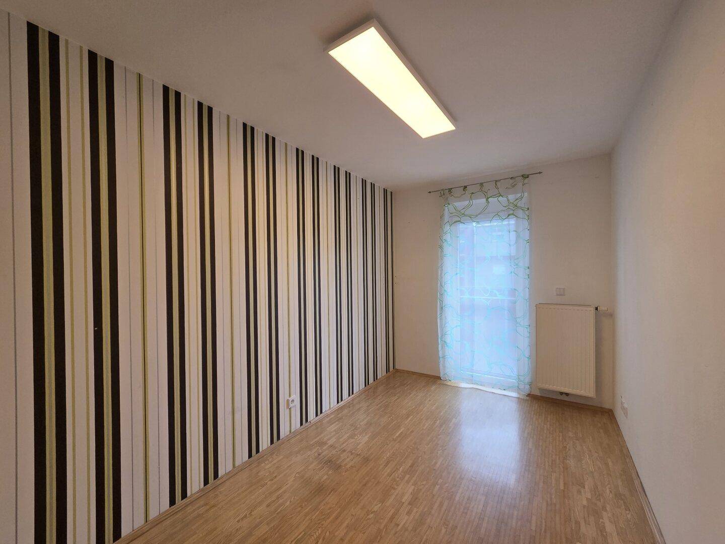 Zimmer mit gestreifter Tapete, Laminatboden und Fenster mit Vorhang.