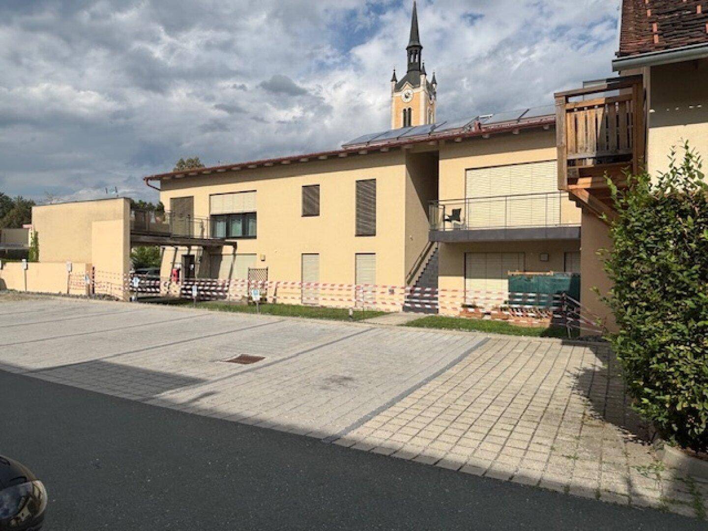Modernes Gebäude mit Parkplatz davor und einem Kirchturm im Hintergrund unter bewölktem Himmel.
