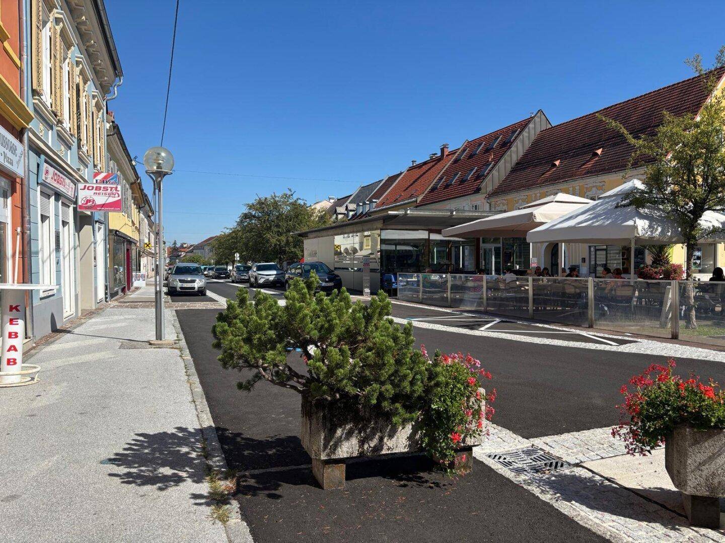 Belebte Straße mit Geschäften, Restaurants und parkenden Autos unter blauem Himmel.