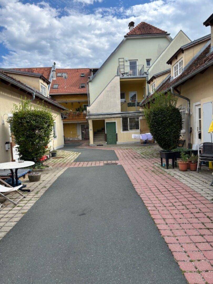 Innenhof zwischen Gebäuden mit gepflasterten Wegen, Pflanzen und Sitzgelegenheiten.