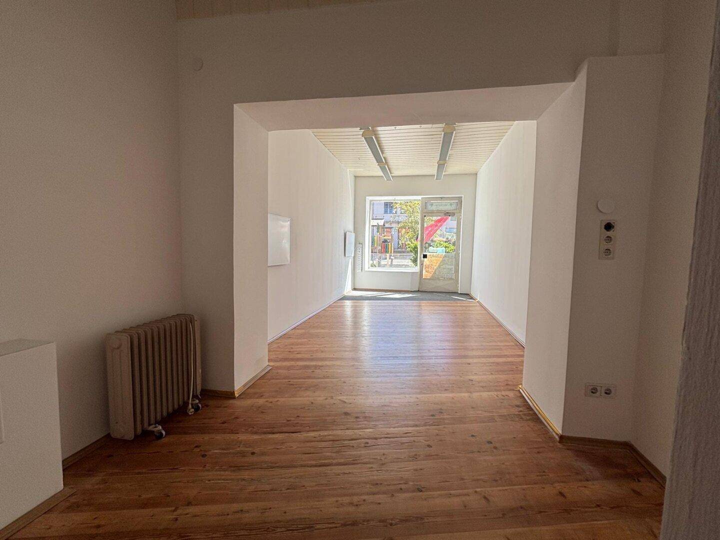 Langer, heller Raum mit Holzboden, Heizkörper und Blick zur Eingangstür und Fenstern.