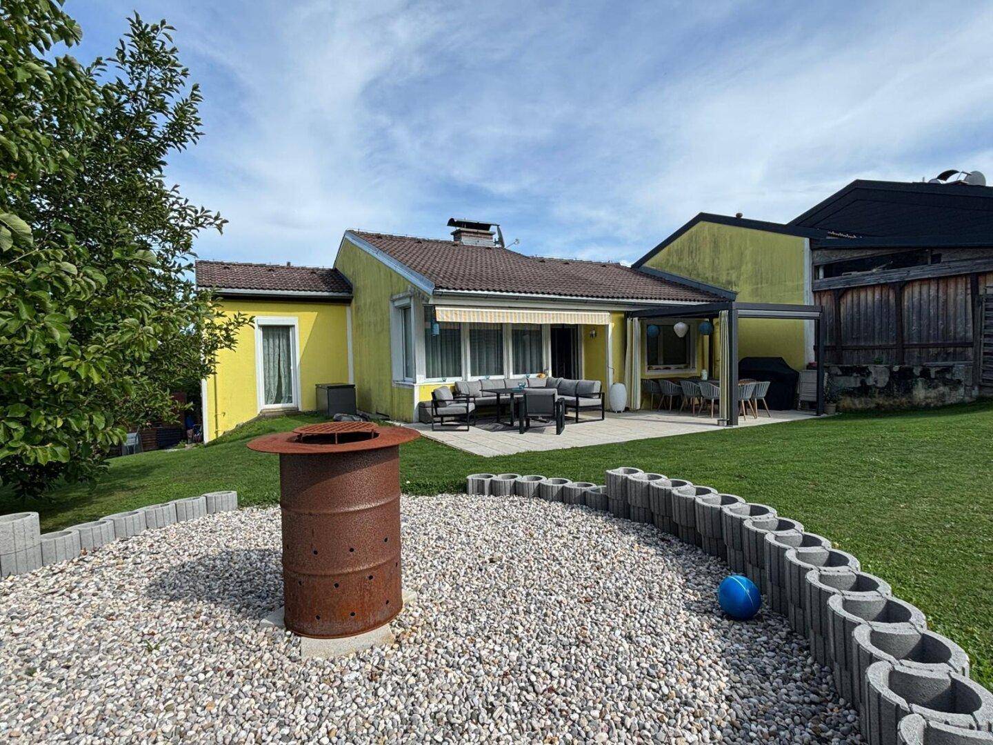 Weitläufiger Garten mit Rasenfläche, Feuerstelle und gepflasterter Terrasse am Haus.