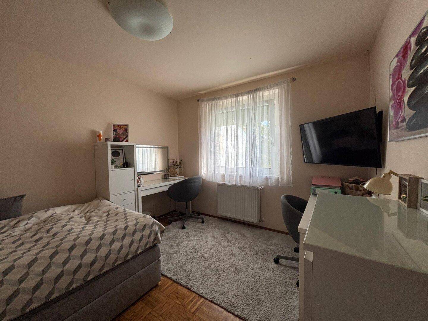 Helles Schlafzimmer mit Fenster, Schreibtisch und Teppichboden unter dem Schreibtisch.