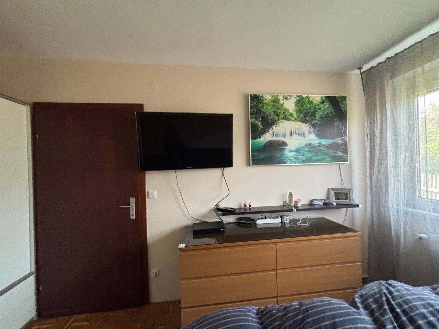 Schlafzimmer mit Fernseher, Kommode und Fenster, das viel Licht hereinlässt.