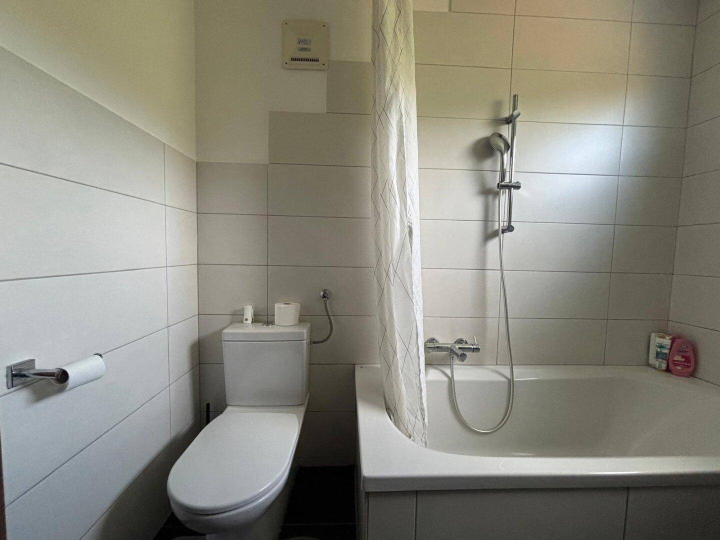 Helles Badezimmer mit Badewanne, Duschvorhang und WC, ausgestattet mit hellen Fliesen.