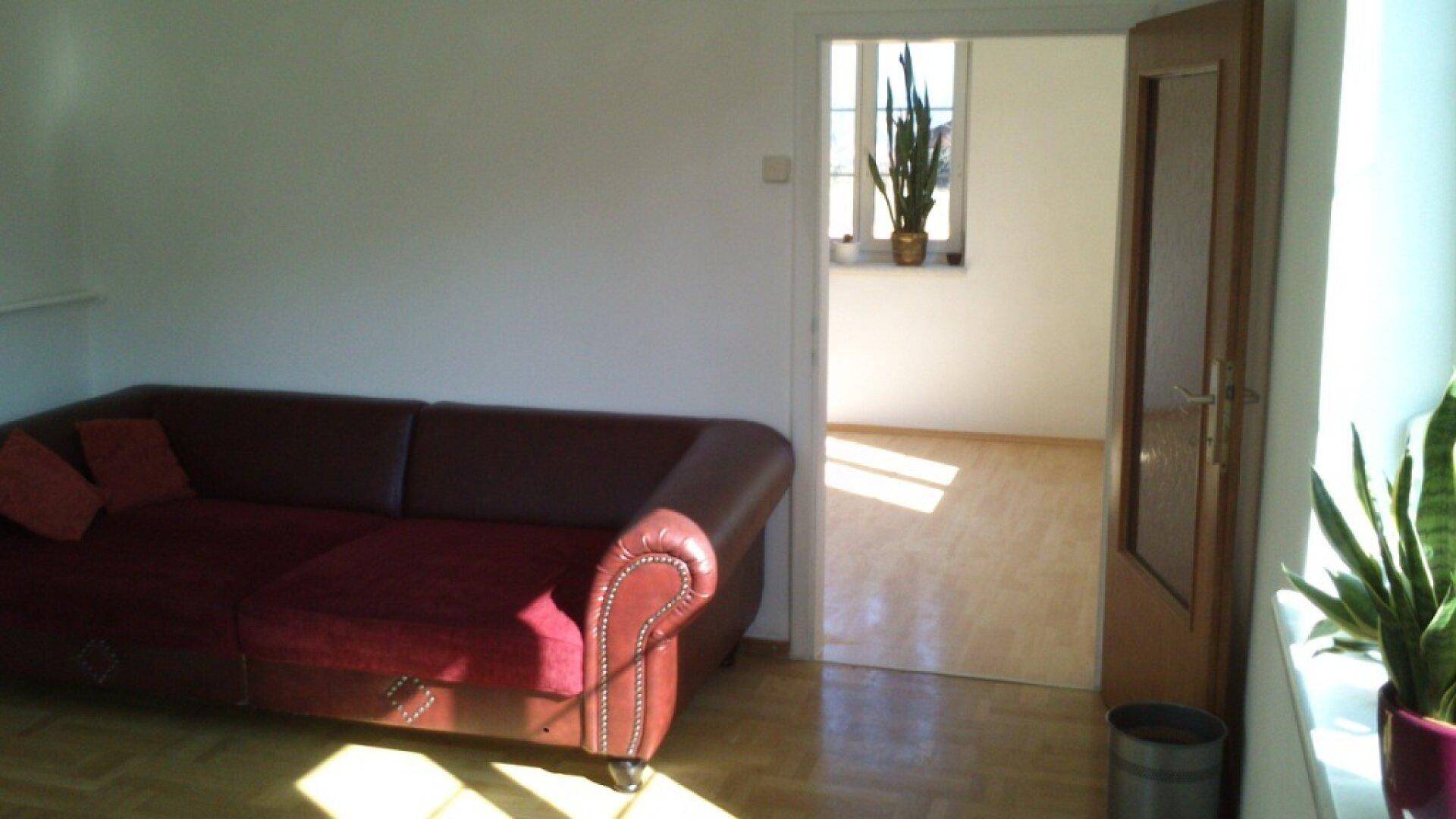Helles Wohnzimmer mit rotem Sofa, Parkettboden und Blick in einen weiteren Raum mit Fenster.