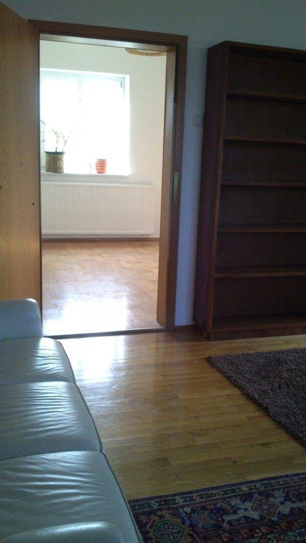 Wohnzimmer mit hellem Sofa, Parkettboden und Blick in einen angrenzenden Raum mit Fenster und Heizung.