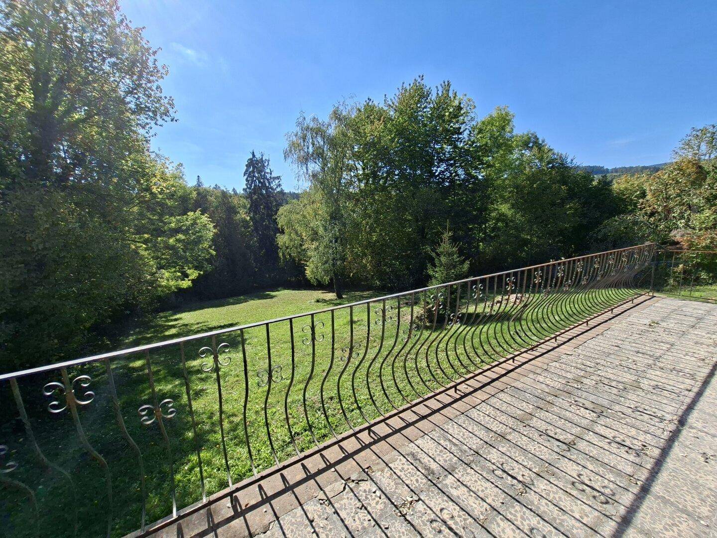 Panoramablick von einem Balkon mit schmiedeeisernem Geländer über eine sonnige Gartenlandschaft.