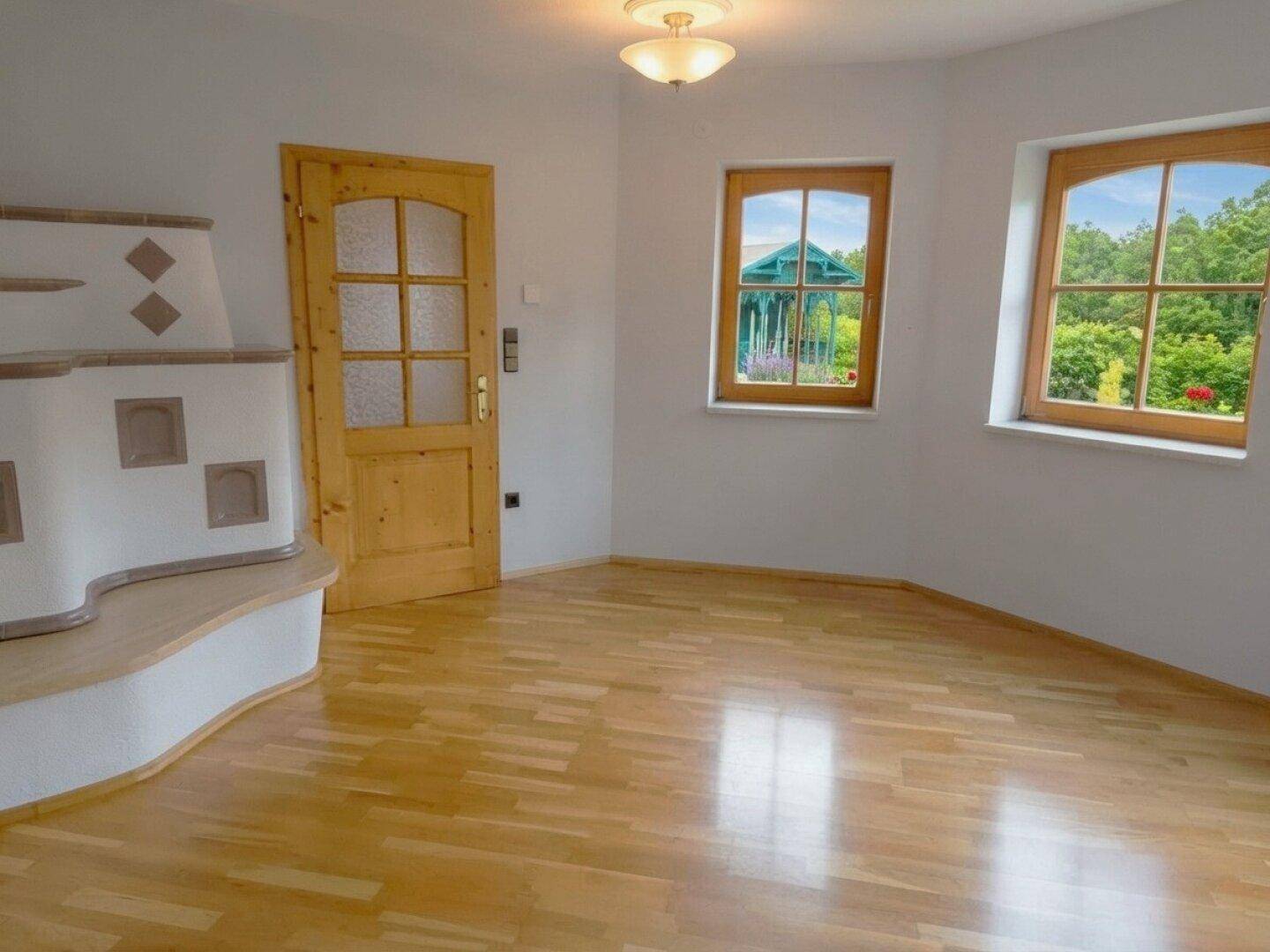 Heller Wohnraum mit Kachelofen, Holzboden und Fenstern mit Blick ins Grüne.
