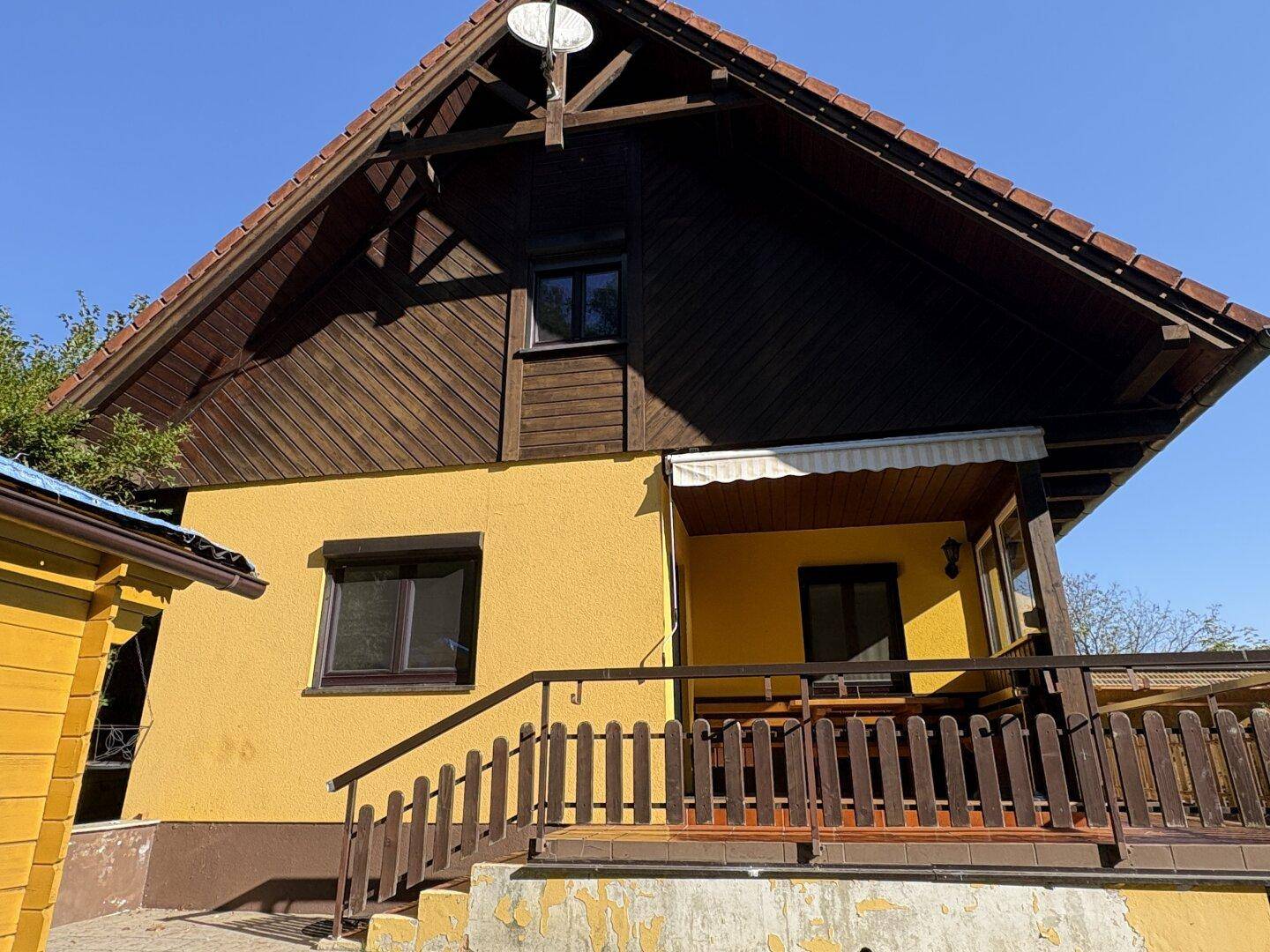 Seitenansicht des Hauses mit gelber Fassade, Holzbalkon und Satteldach unter blauem Himmel.