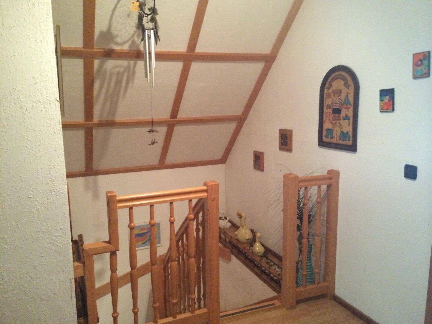 Holztreppe mit Geländer führt in den oberen Bereich, umgeben von weiß gestrichenen Wänden und Dachschrägen.