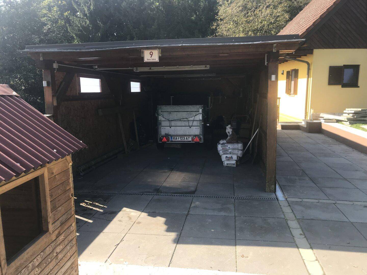 Offener Carport mit gepflastertem Vorplatz und einem Anhänger, neben dem Wohnhaus gelegen.