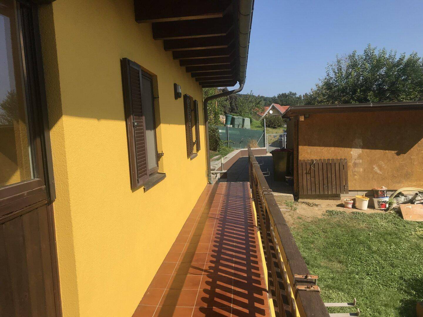 Langer, gefliester Balkon entlang der gelben Hausfassade mit Holzbalkongeländer und Blick ins Grüne.