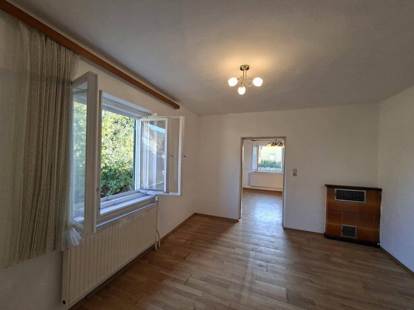 Helles Wohnzimmer mit Holzparkett, großem Fenster und einem traditionellen Kachelofen.