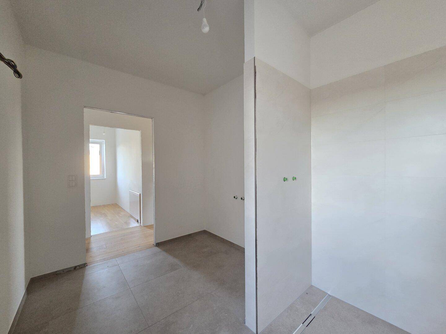 Badezimmer im Rohbau mit grauen Fliesen und vorbereiteten Anschlüssen für Sanitäreinrichtungen.