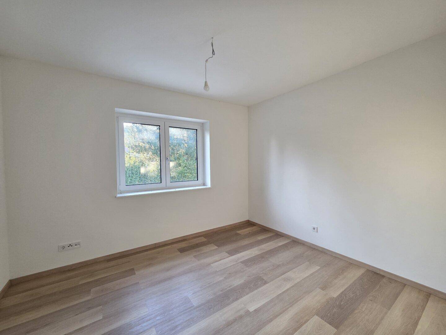 Leeres Schlafzimmer mit Laminatboden und einem Fenster, das viel Tageslicht hereinlässt.