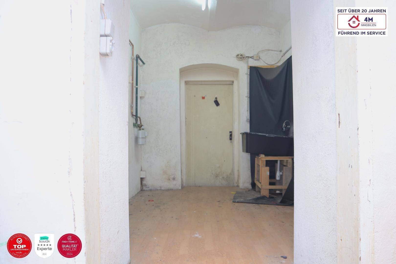 Schmaler Flur mit einer Tür am Ende und sichtbaren Versorgungsleitungen an der Wand, in einfachem Zustand.