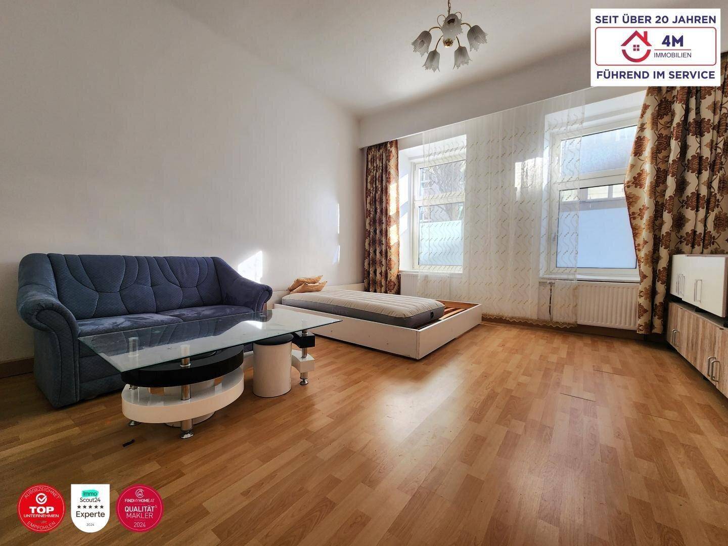 Wohnbereich mit Laminatboden, Sofa und zwei Fenstern, die viel Tageslicht hereinlassen.