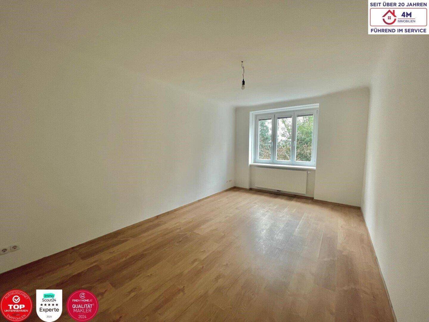 Leeres Zimmer mit hellem Holzboden und einem Fenster mit Blick ins Grüne.