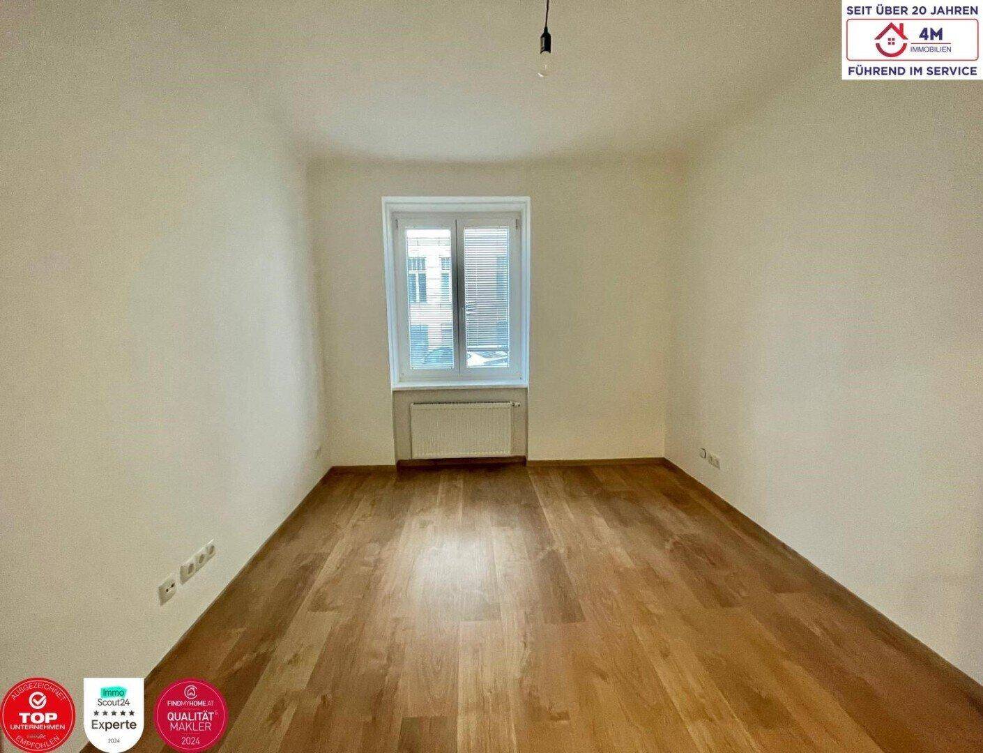 Helles Schlafzimmer mit Holzboden und einem Fenster mit Blick auf die umliegenden Gebäude.