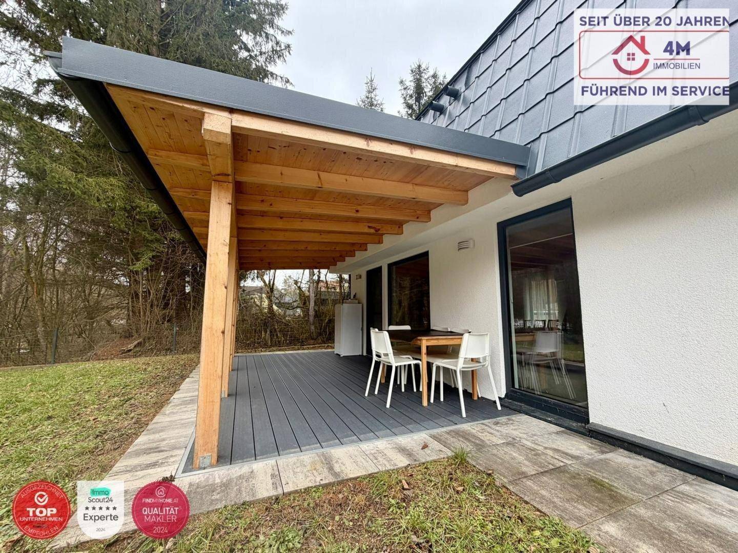 Überdachte Holzterrasse mit Esstisch und Stühlen, umgeben von Grünflächen.