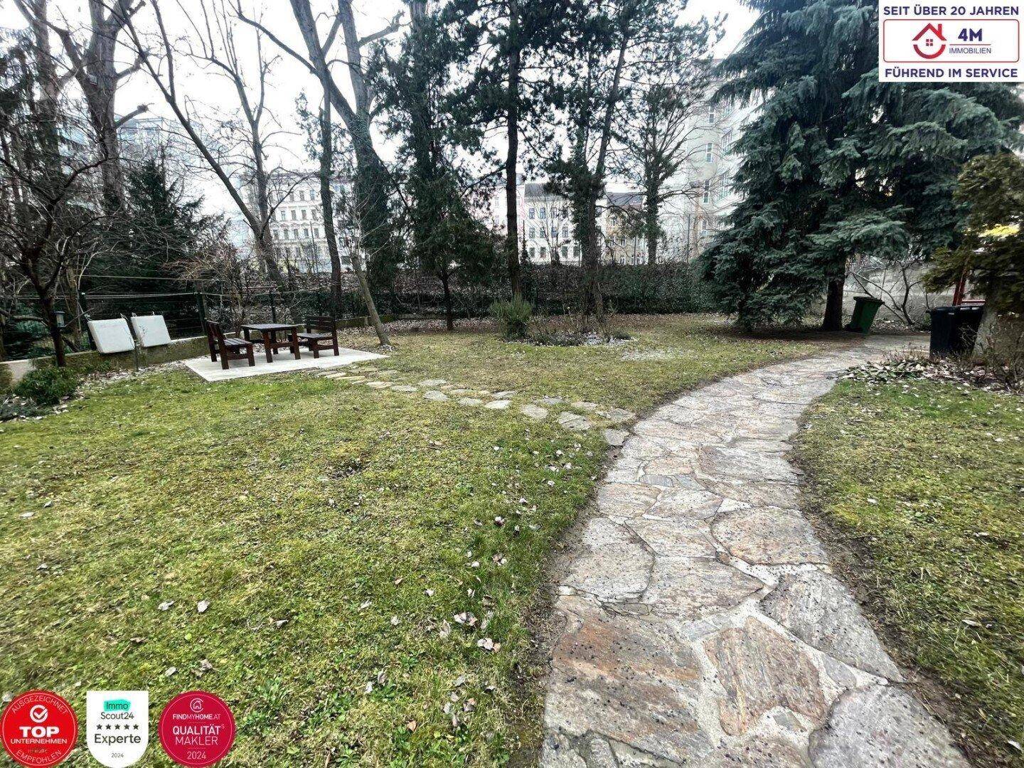 Großer Gemeinschaftsgarten mit Steinpfad, Bäumen und einem Sitzbereich im Freien.