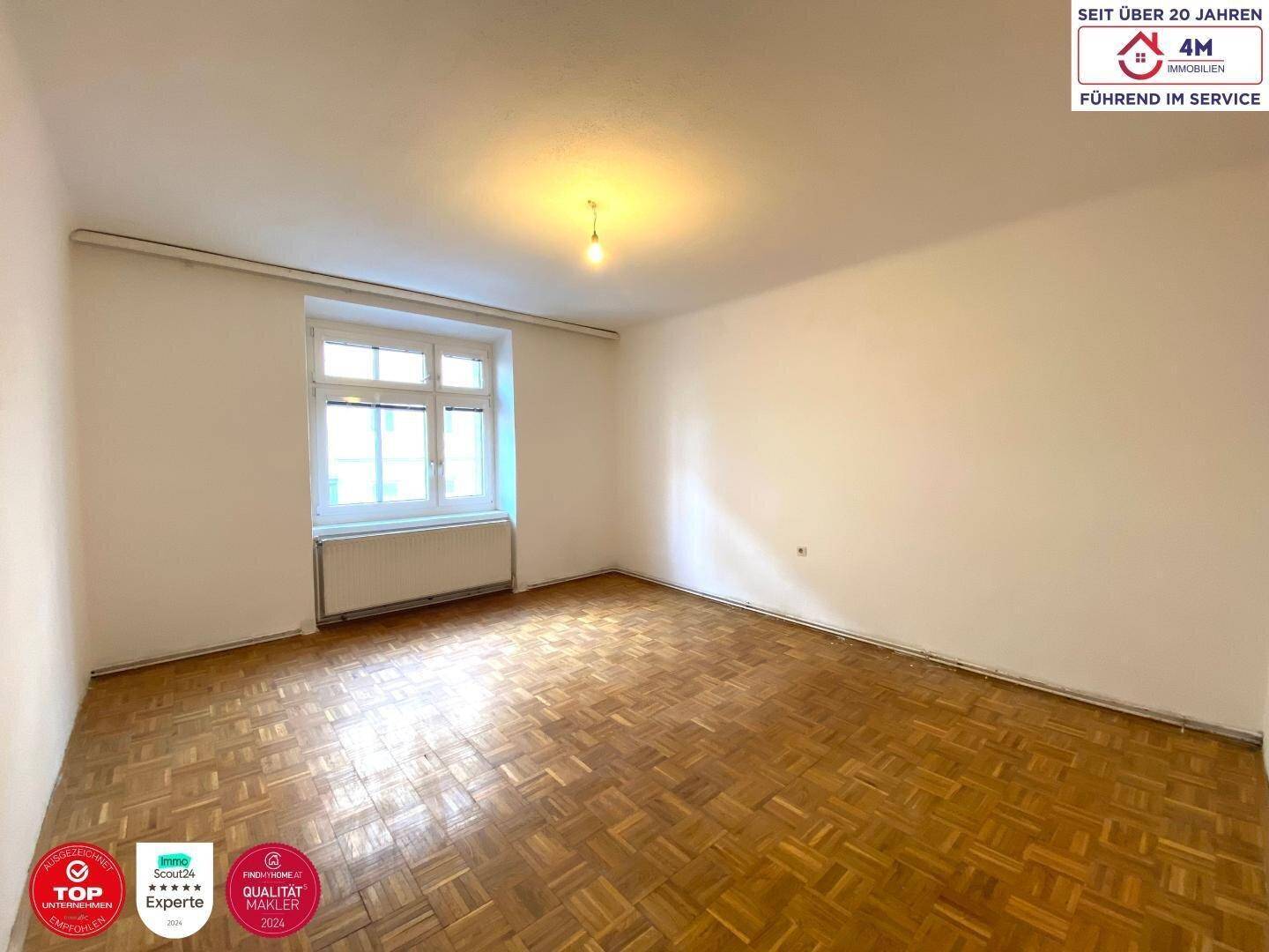 Helles, leeres Zimmer mit Fischgrätparkett und großem Fenster, ideal als Wohn- oder Schlafzimmer.