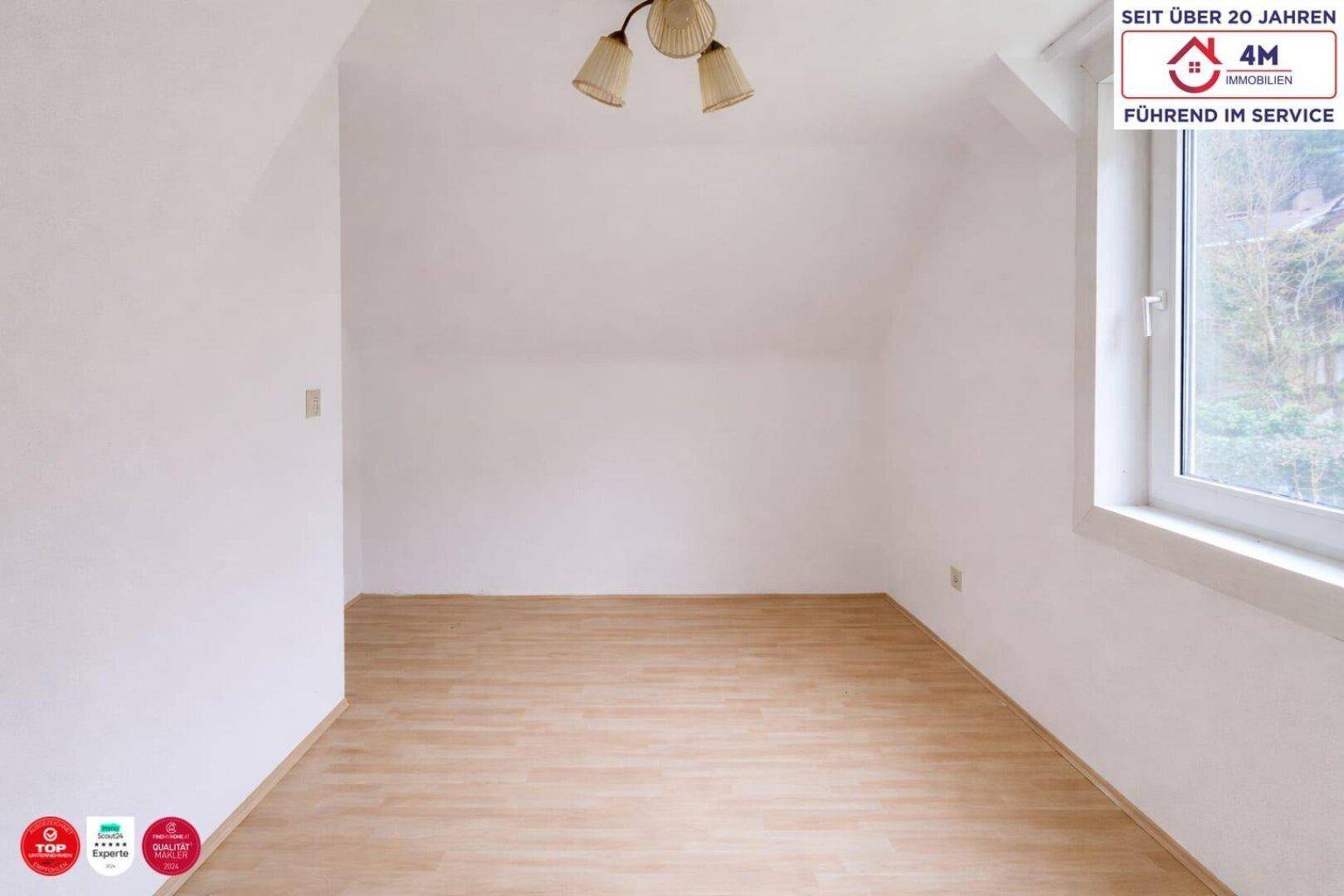 Helles Dachzimmer mit schrägen Wänden, Laminatboden und einem Fenster mit Ausblick.