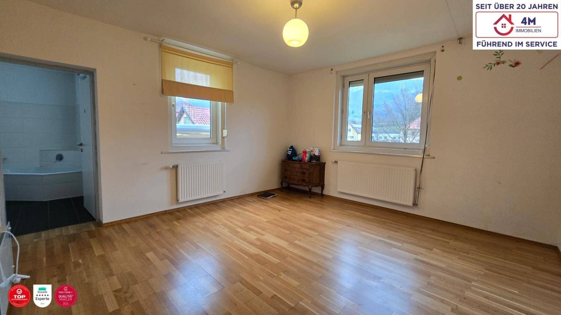 Schlafzimmer mit Holzboden und zwei Fenstern, angrenzend an ein Badezimmer.