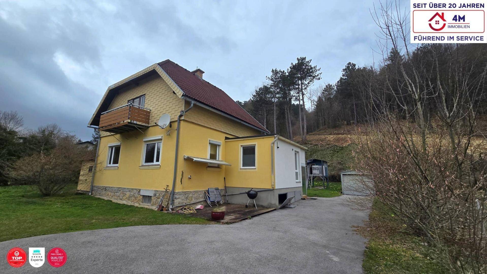 Gelbes Einfamilienhaus mit Satteldach, Balkon und Anbau, umgeben von Bäumen und Natur.