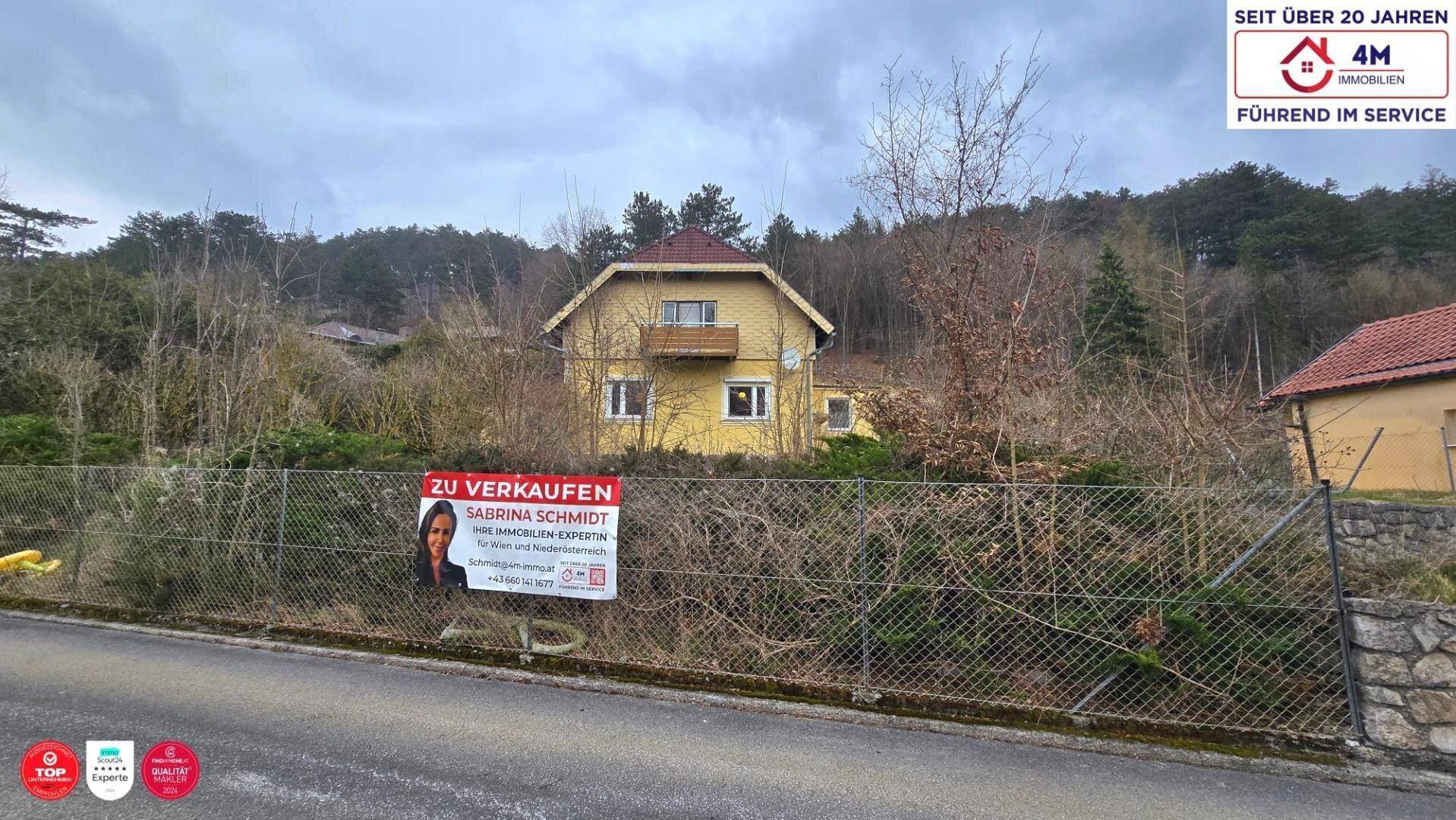 Freistehendes gelbes Haus in ländlicher Umgebung, sichtbar hinter einem Zaun mit Verkaufsplakat.
