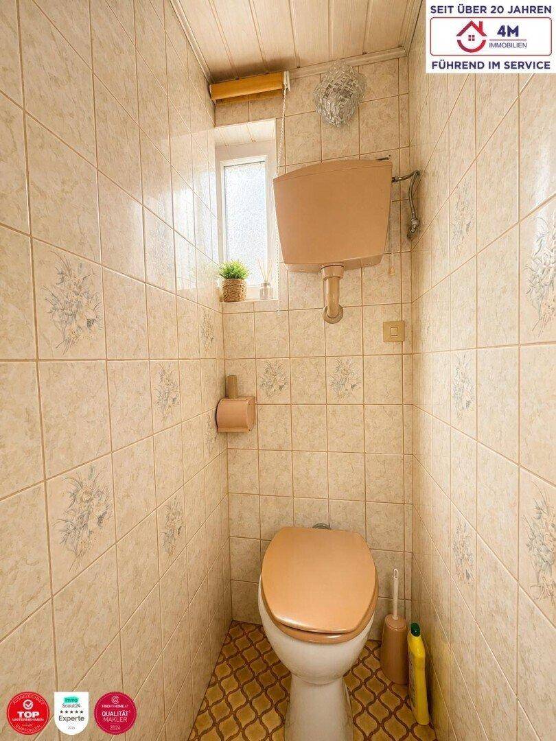 Kleines WC mit beigefarbenen Fliesen, Fenster und einer Holzdecke.