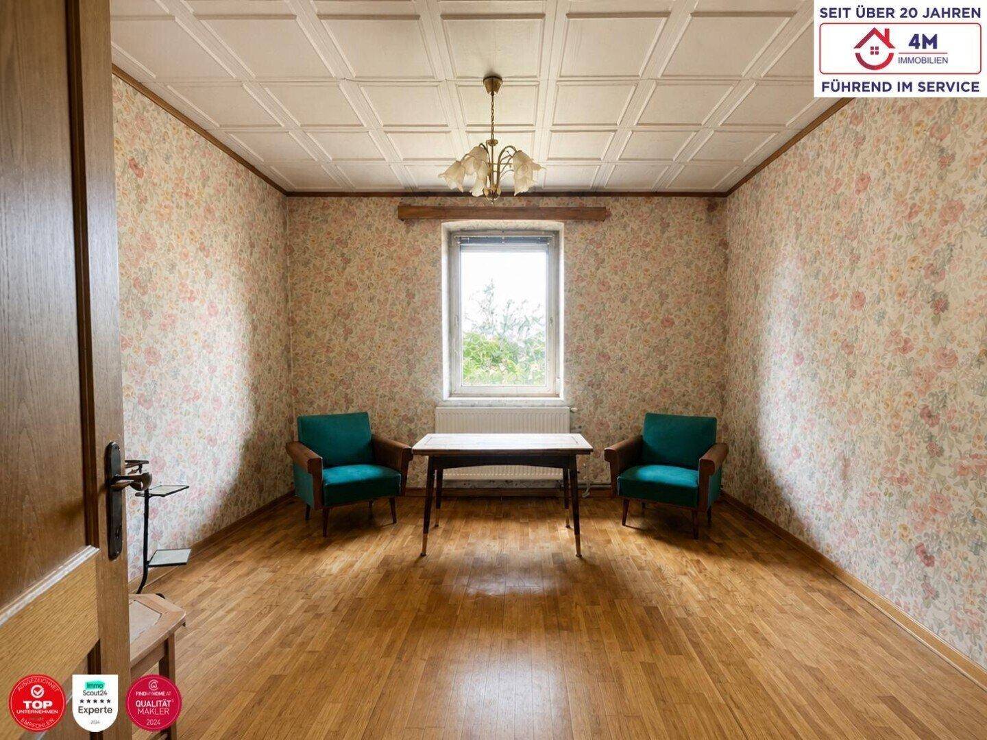 Wohnzimmer mit floraler Tapete, Holzboden und zwei Vintage-Sesseln unter einer Kassettendecke.