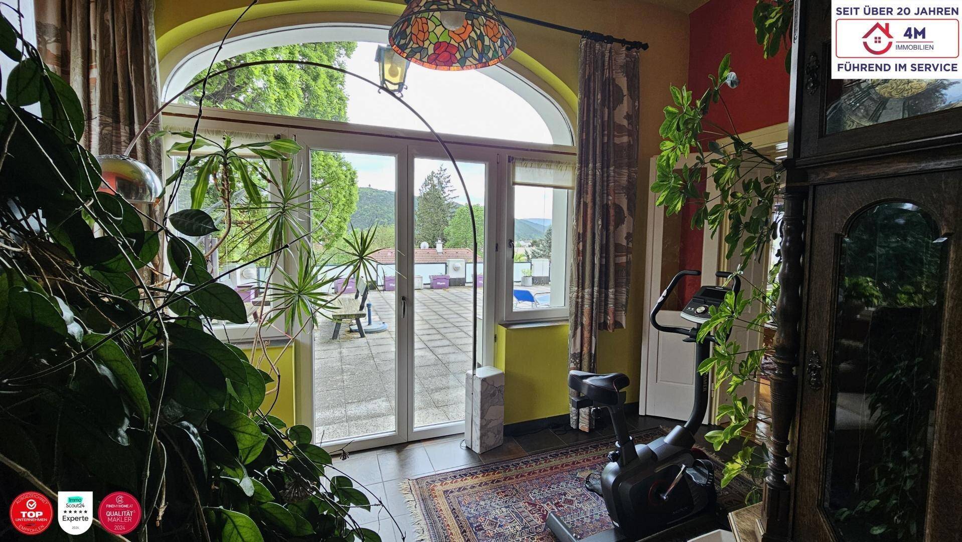 Heller Raum mit großen Bogenfenstern, die zur Terrasse führen, ausgestattet mit einem Heimtrainer und Pflanzen.