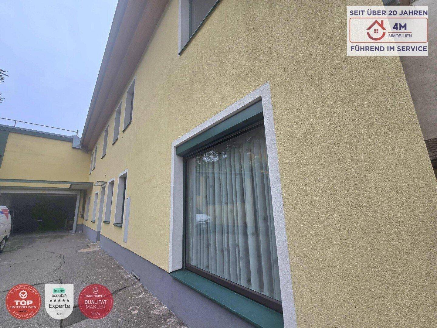 Gelbe Gebäudefassade mit einem Fenster, das einen Rollladen besitzt, und einem Carport im Hintergrund.
