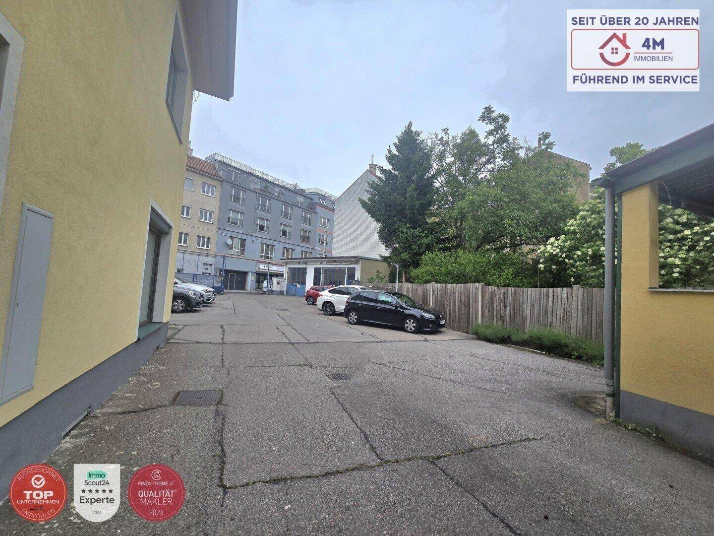Außenbereich mit Parkplätzen und Blick auf weitere Gebäude in der Ferne, umgeben von Bäumen.