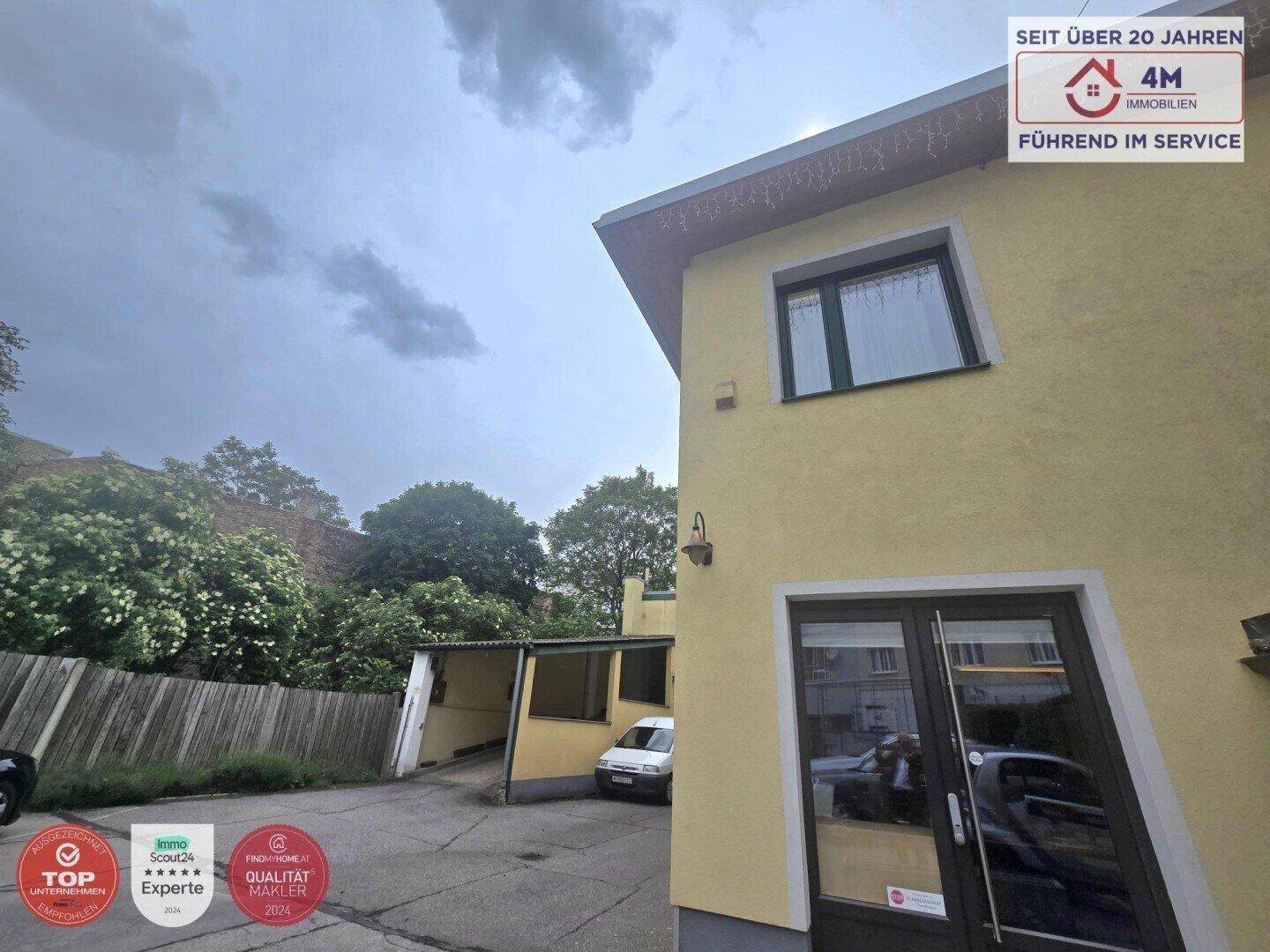 Gelbe Gebäudefassade mit dunklen Fenstern und einer Doppeltür, umgeben von Bäumen und einem Carport.