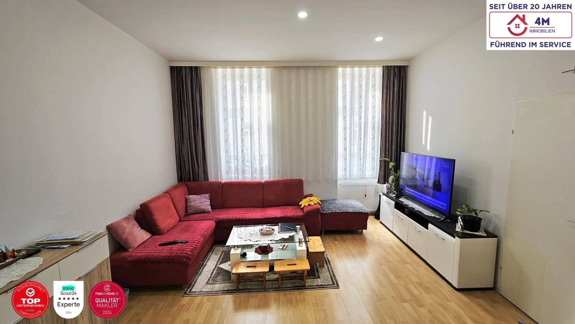 Gemütliches Wohnzimmer mit großem roten Ecksofa, Holzboden und hellen Fenstern mit Vorhängen.