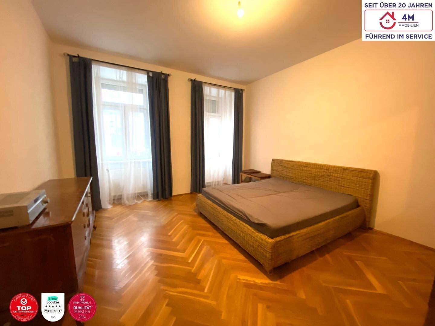 Schlafzimmer mit Fischgrätparkett, Doppelbett und zwei Fenstern.