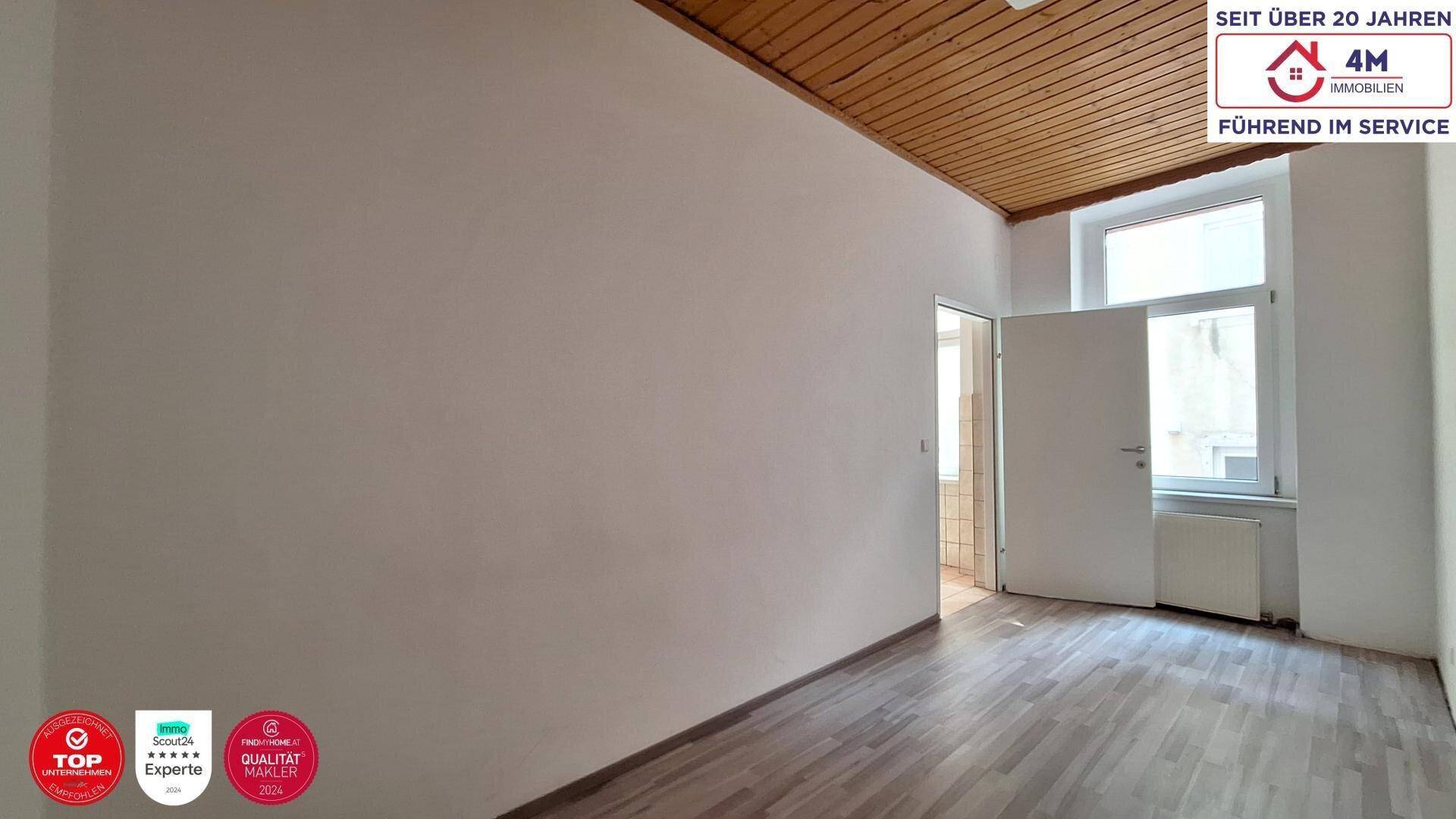 Helles Zimmer mit Holzbalkendecke, Laminatboden und Zugang zu einem weiteren Raum, gut belichtet.