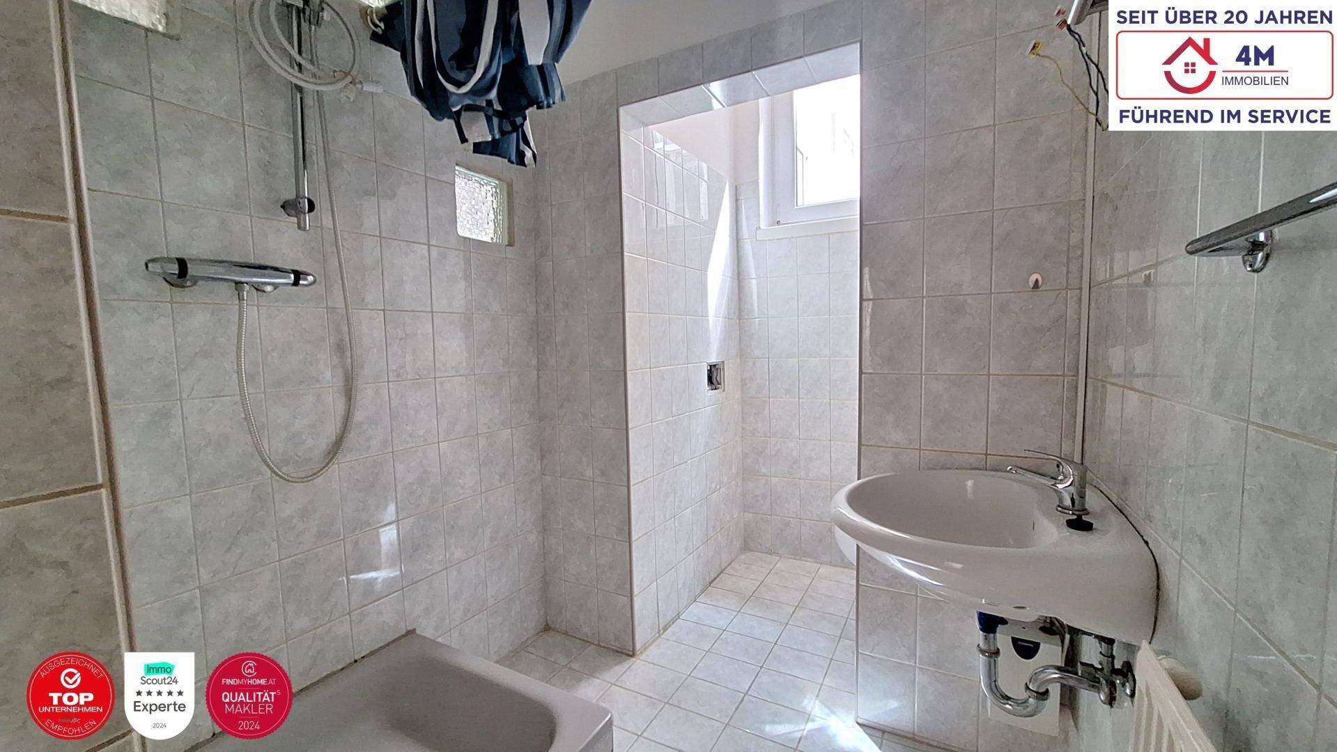 Gefliestes Badezimmer mit Dusche, Waschbecken und einem Fenster für natürliche Belüftung und Tageslicht.