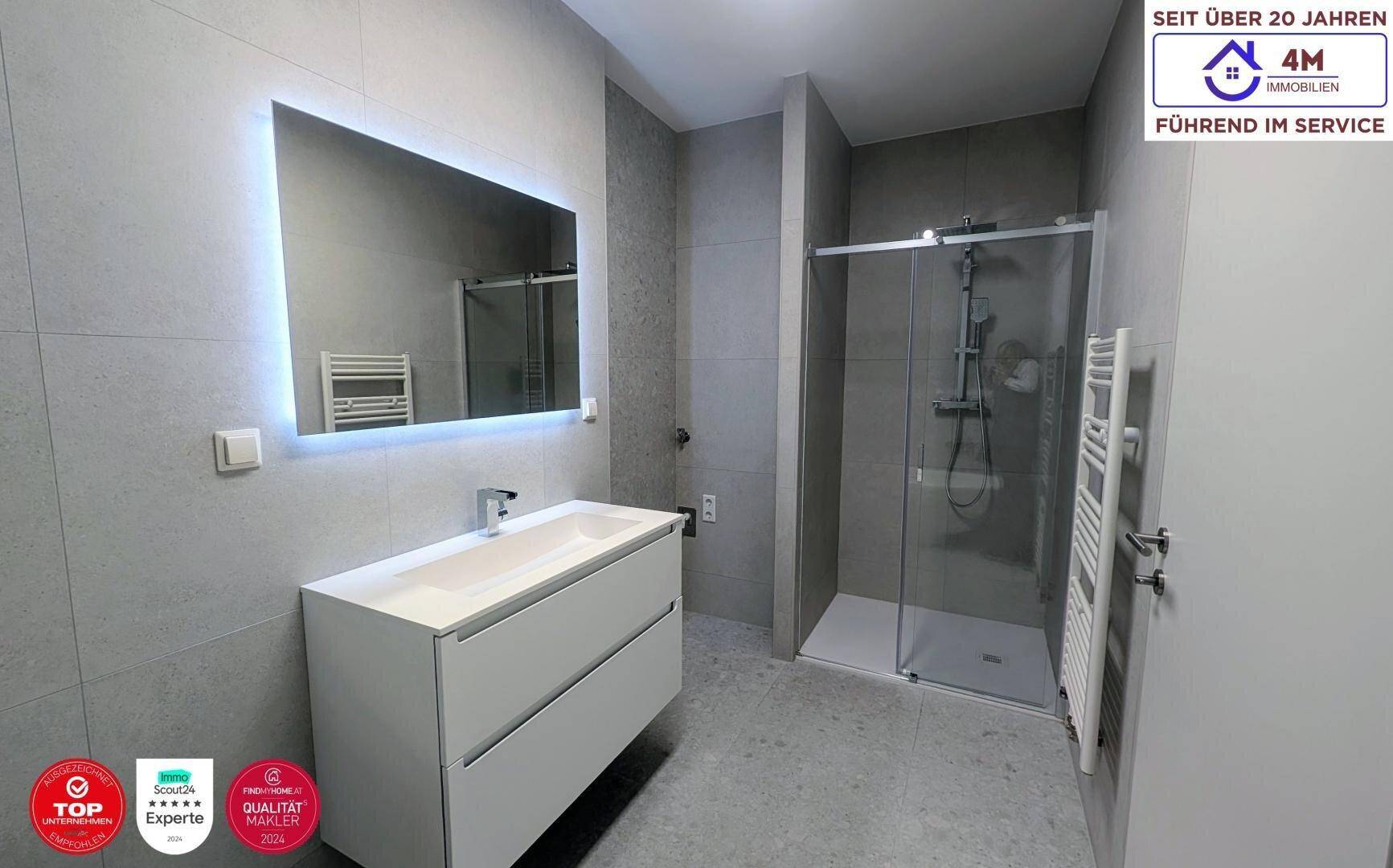 Modernes Badezimmer mit grauen Fliesen, beleuchtetem Spiegel, Einzelwaschbecken und begehbarer Dusche.