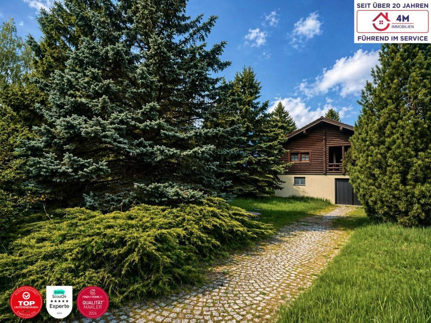 Außenansicht des Blockhauses, umgeben von hohen Nadelbäumen und einer gepflasterten Zufahrt.