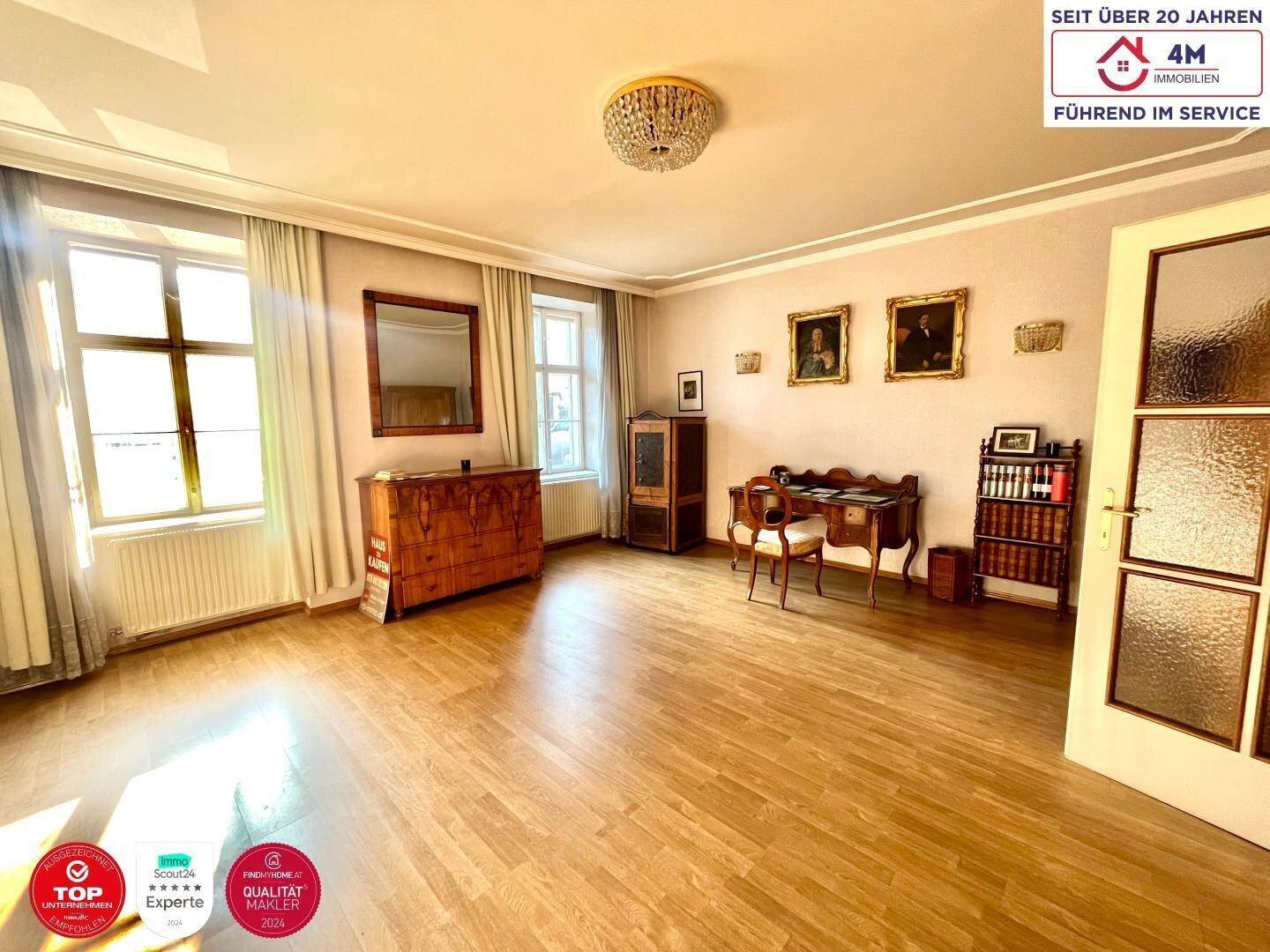 Geräumiges Wohnzimmer mit Holzboden, klassischen Möbeln und viel Tageslicht durch große Fenster.