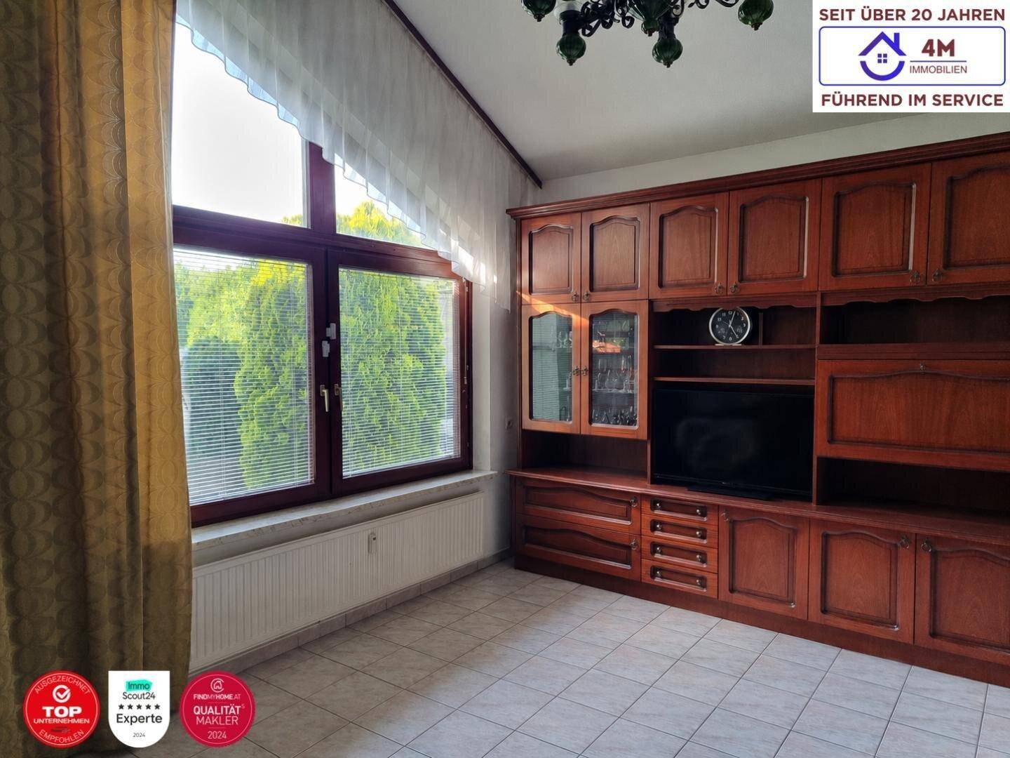 Wohnzimmer mit großem Holzschrank, Fliesenboden und Fenster mit Blick ins Grüne.