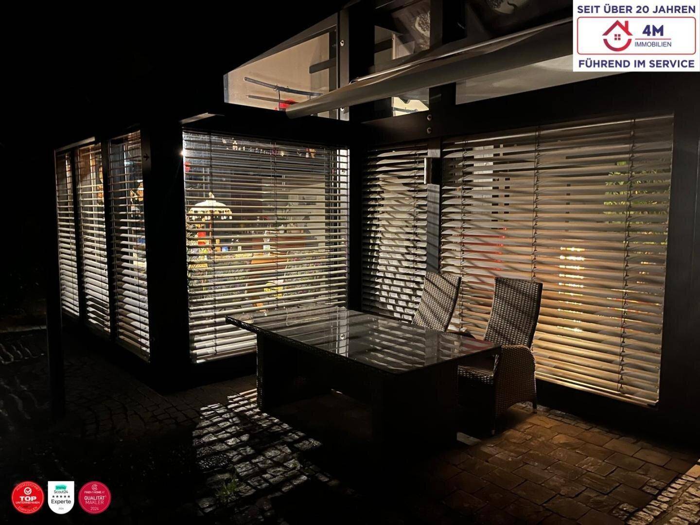 Außenbereich bei Nacht mit einem Glastisch und Stühlen auf der Terrasse, beleuchtet von innen.