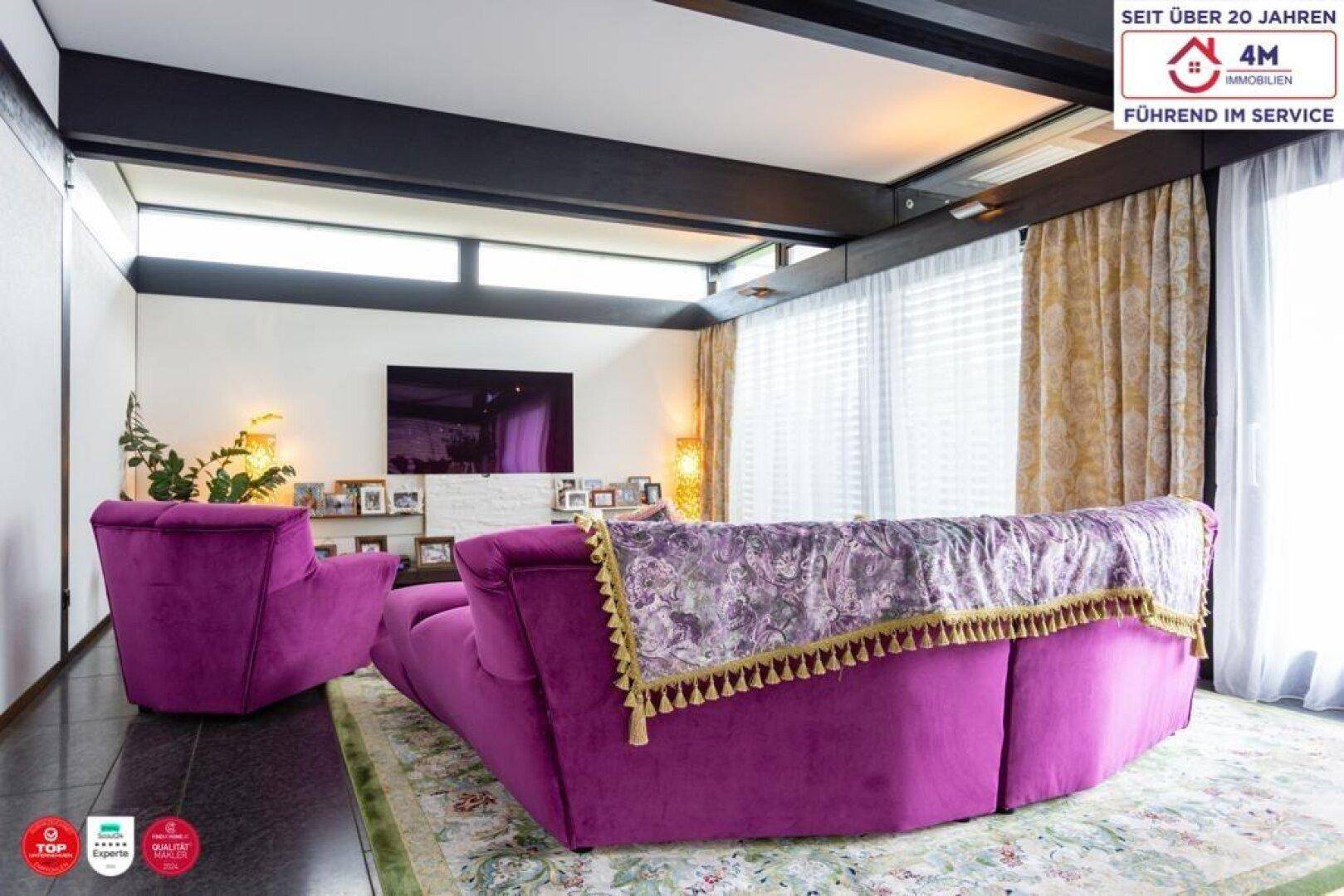 Gemütliches Wohnzimmer mit lila Sofa, großem Fernseher und hellen Vorhängen.