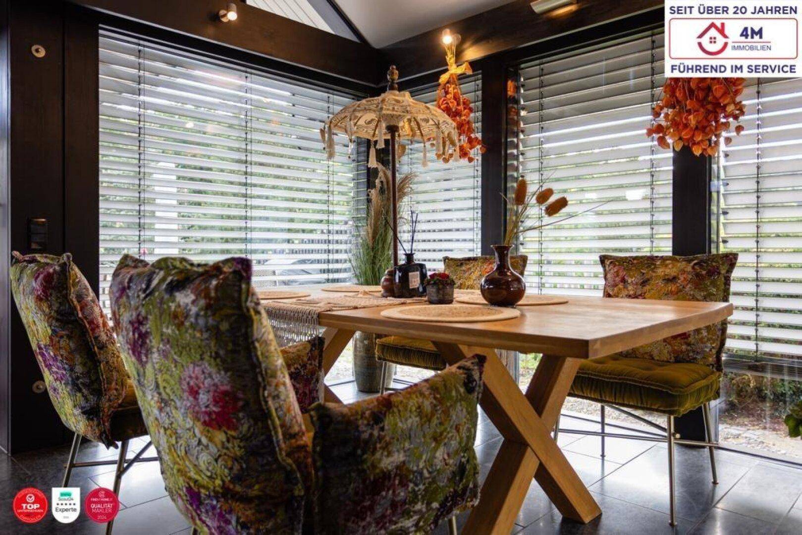 Gemütlicher Essbereich mit Holztisch, floral gemusterten Stühlen und Jalousien an den großen Fenstern.